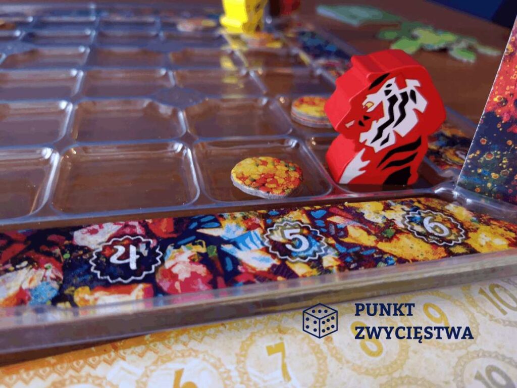 Najniższy poziom gry Holi widoczny czerwony pion w kształcie tygrysa oraz żetony słodyczy w tle fragment żółtego pionka